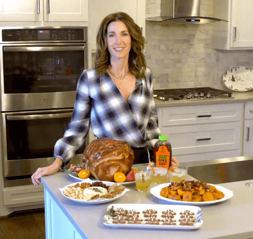 A woman in a kitchen presenting a festive meal including a glazed ham, sides, cookies and a bottle of Nate’s honey.