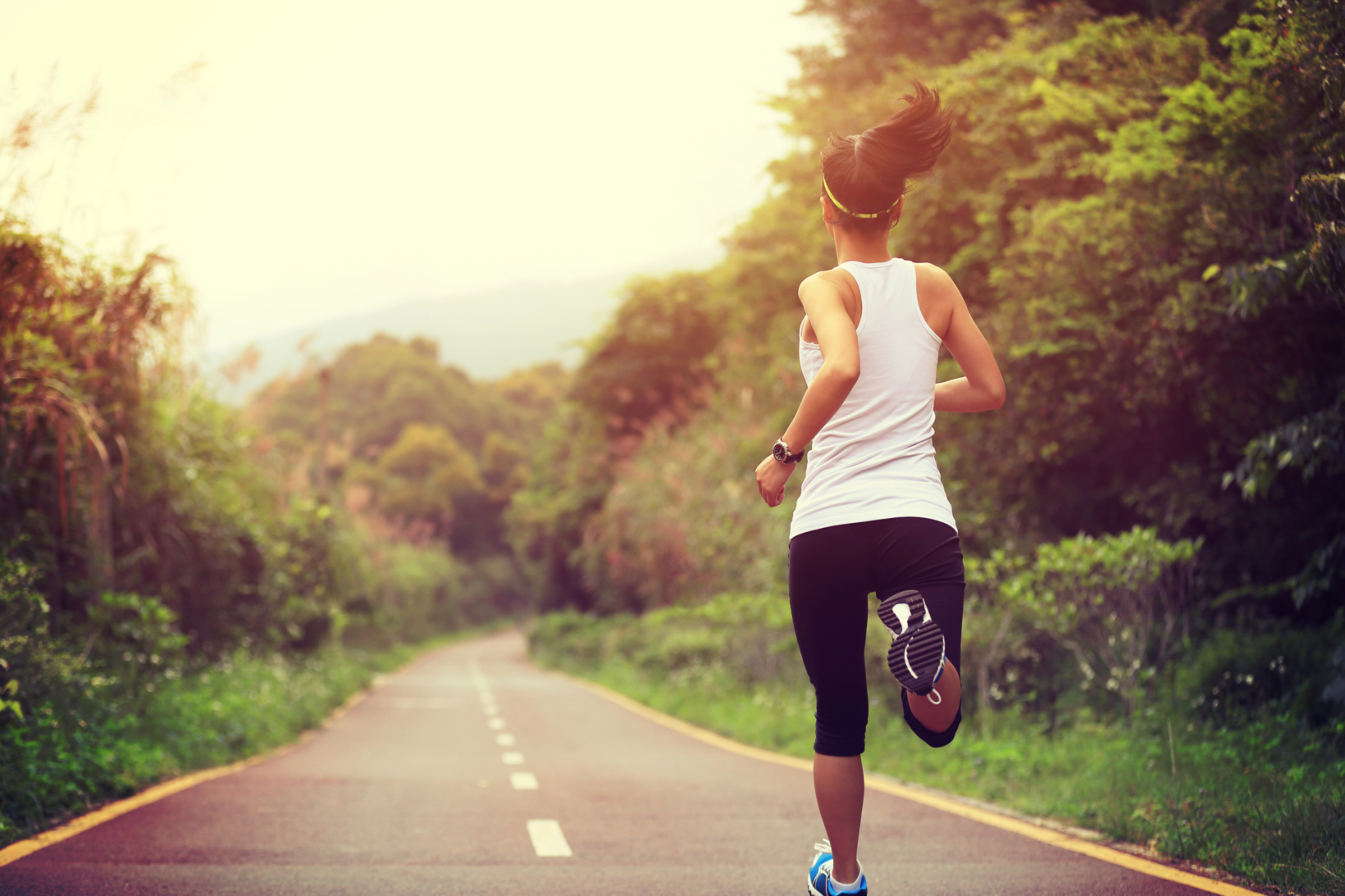 A woman in a white tank top and dark shorts is running along a road, surrounded by lush greenery.