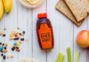 Nate's honey bottle surrounded by snacks, fruits, bread and celery on a wooden background.