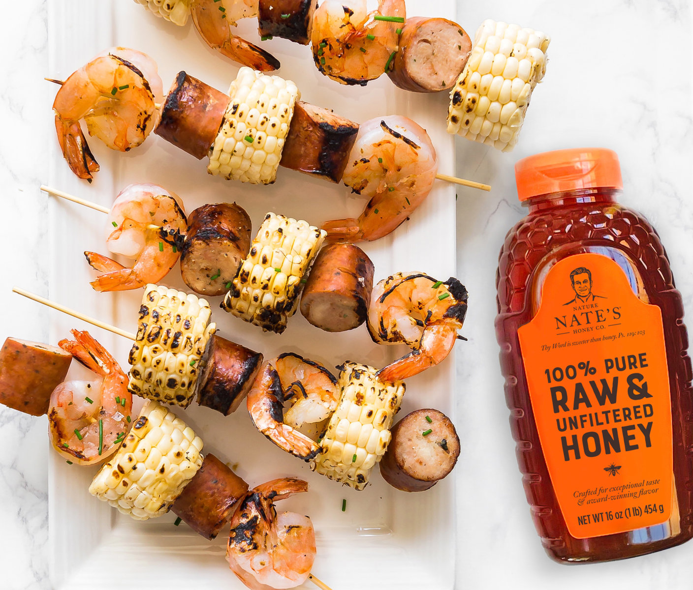 Grilled shrimp, sausage and vegetable skewers with a bottle of Nate's honey on a marble background.