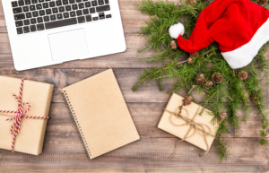 Christmas setting with gifts, Santa hat, and greenery on a wooden surface, alongside a laptop and notepad.