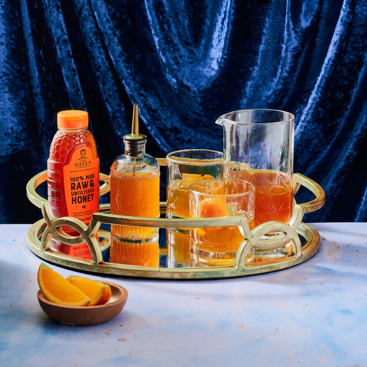 Brass tray with a bottle of Nate's honey, a pitcher and two glasses filled with cocktails and a bottle of simple syrup.