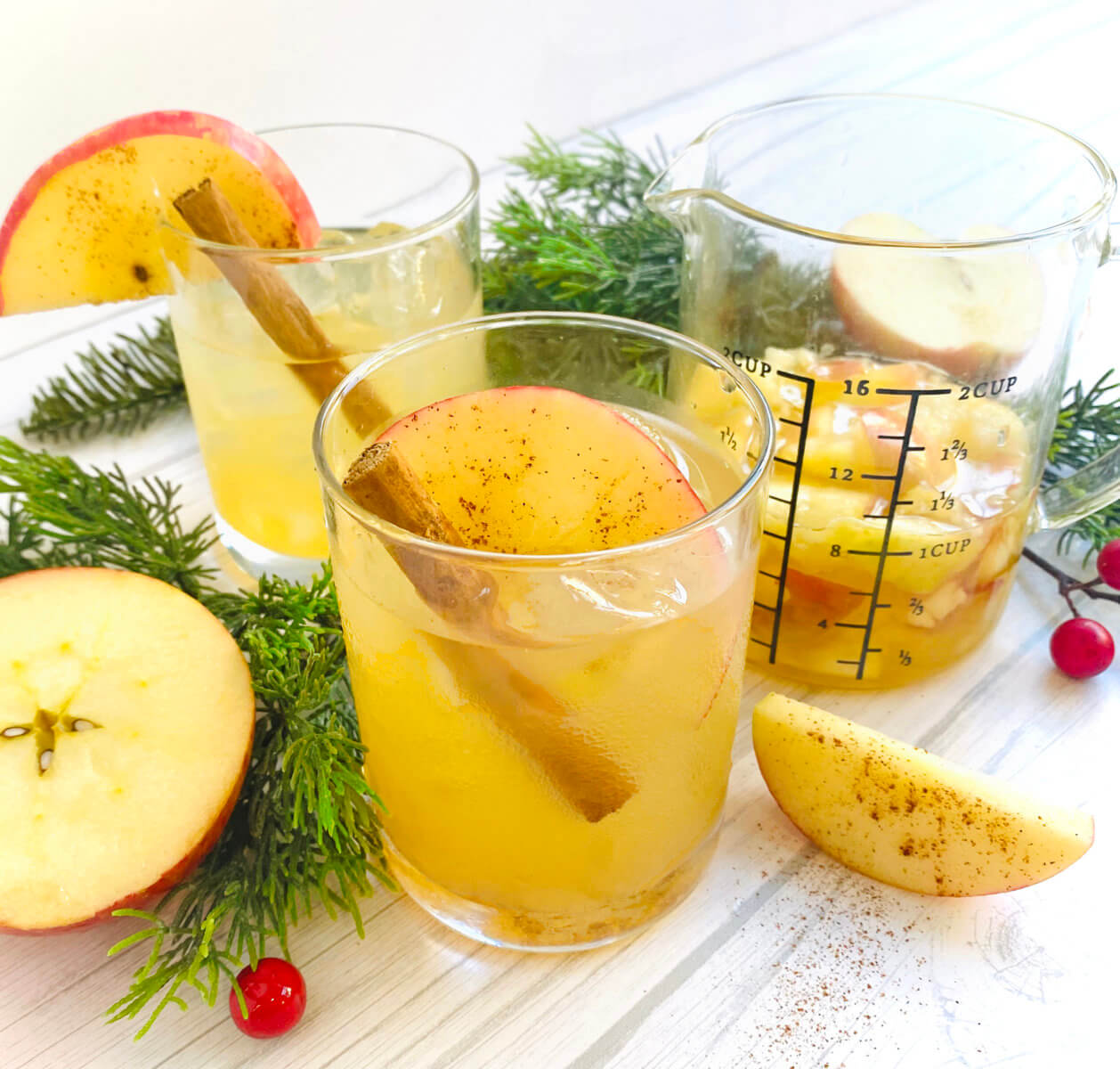 Honey apple cocktails in clear glasses, garnished with apples and cinnamon, surrounded by fresh greenery and berries.