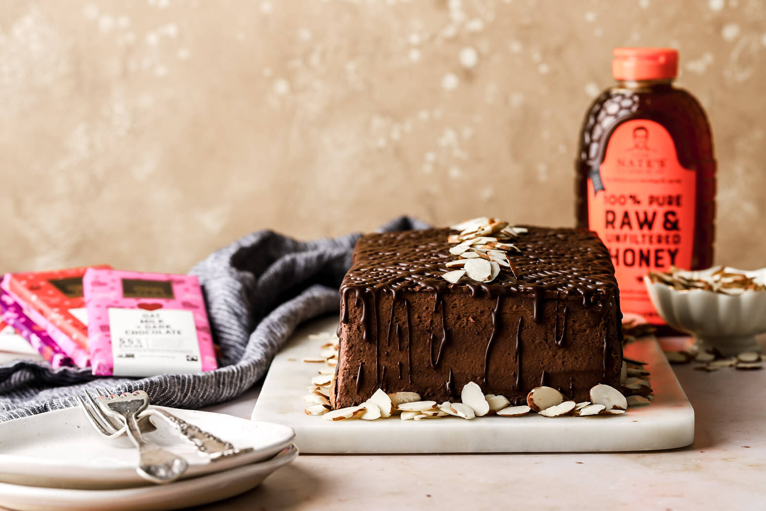 Chocolate cake with almond slices and a Nate's honey bottle, next to a grey napkin on a white marble counter.