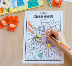 Child's hand coloring a bee on a Color by Numbers activity sheet with crayons and Nate's honey minis nearby. Child's hand coloring a bee on a Color by Numbers activity sheet with crayons and Nate's honey minis nearby.