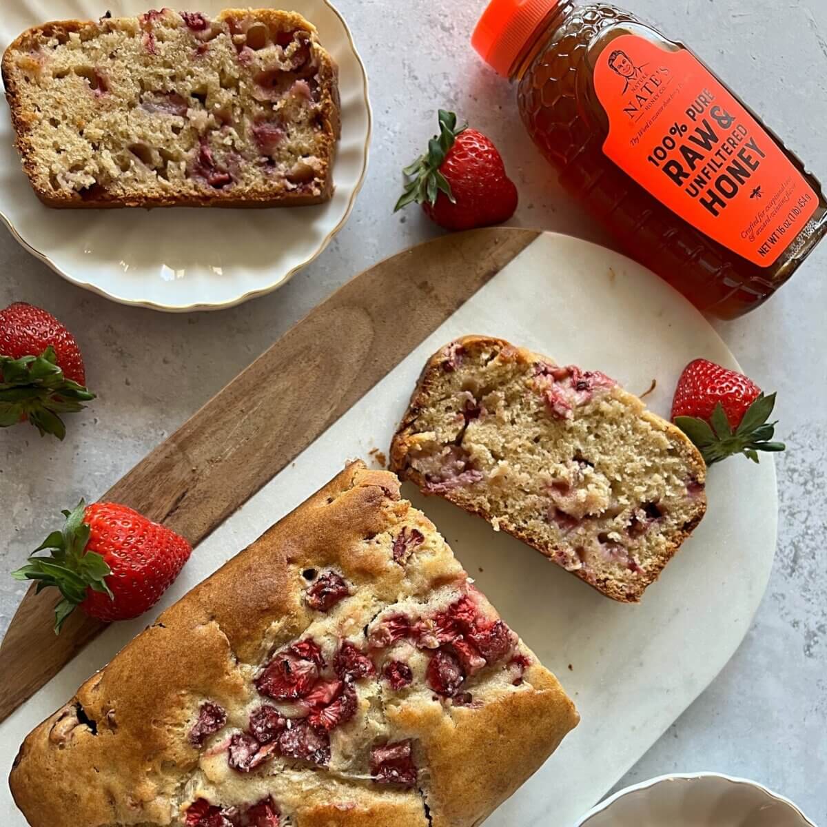 Sliced strawberry bread cake with a bottle of Nate’s honey on a marble countertop.