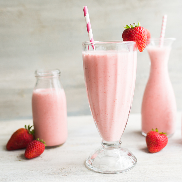 Strawberry smoothies in glass and bottle with straws and fresh berries on a light backdrop.