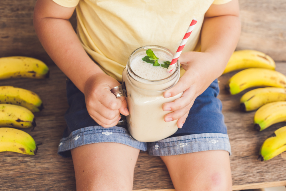 Child holding a banana smoothie with bananas on a wooden table.