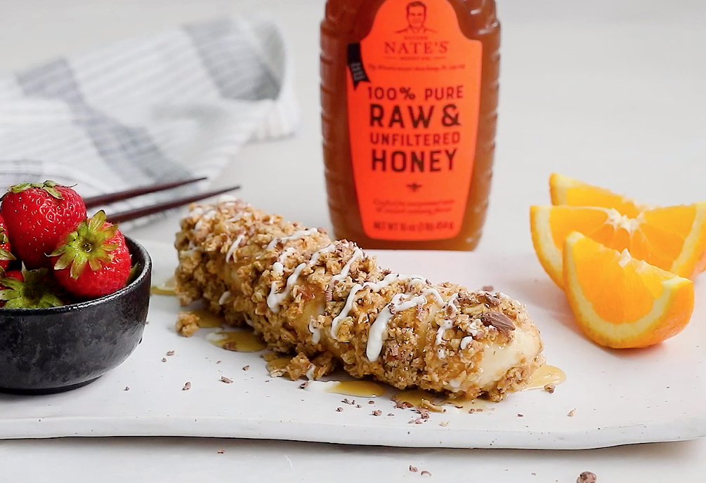 A granola bar drizzled with honey, fresh strawberries, orange slices, and a bottle of Nate's Honey.