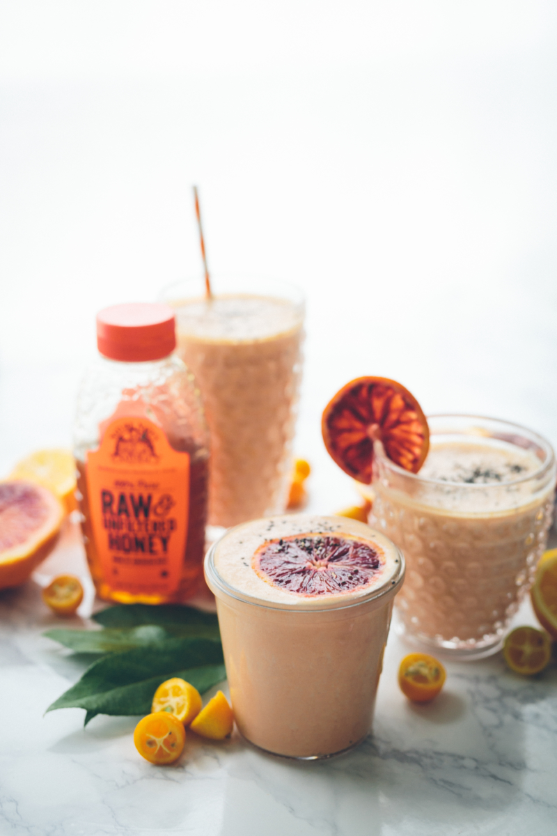 Sweet potato and orange smoothies with a bottle of Nate's raw honey and fresh fruits on a marble surface.