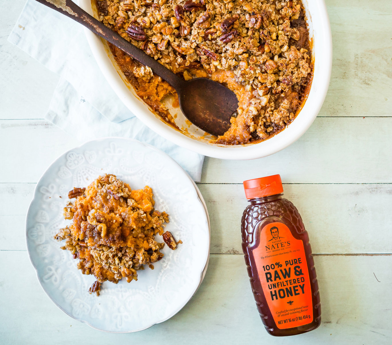 Baked sweet potato casserole in a baking dish and served on a plate and a bottle of Nate's honey on a wooden surface.