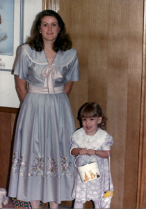 A mother and young daughter dressed in vintage attire, with the child holding a purse, capture a moment from a bygone era.