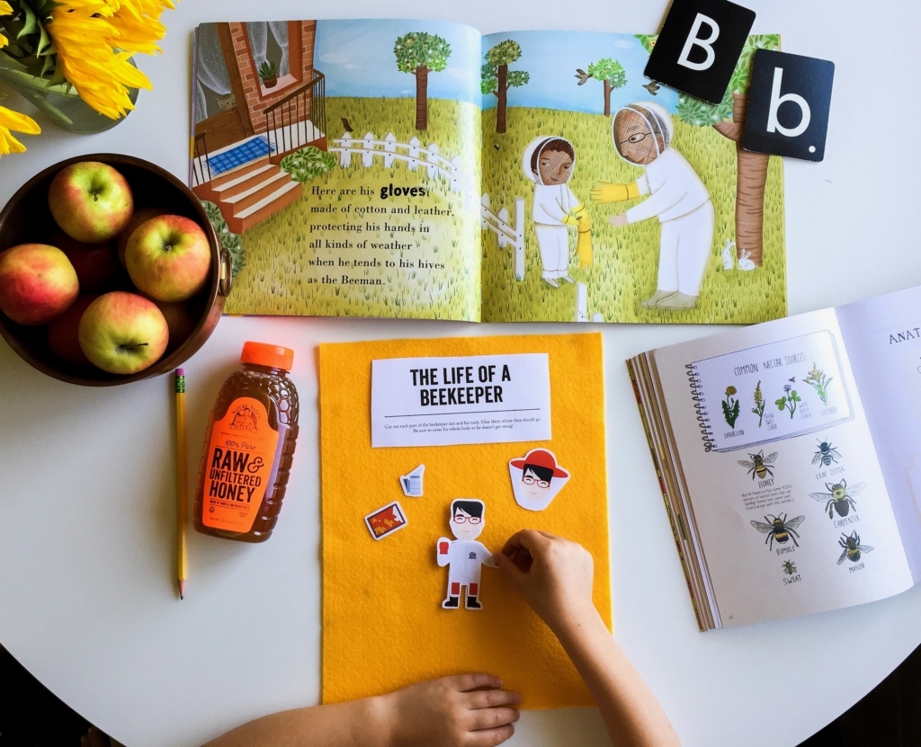 Child's hands with beekeeping books, Nate's honey bottle, apples, sunflowers on a white table.