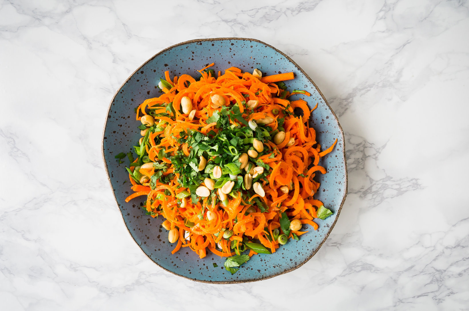 Spiralized carrot salad with peanuts and herbs on a speckled blue plate, marble background.