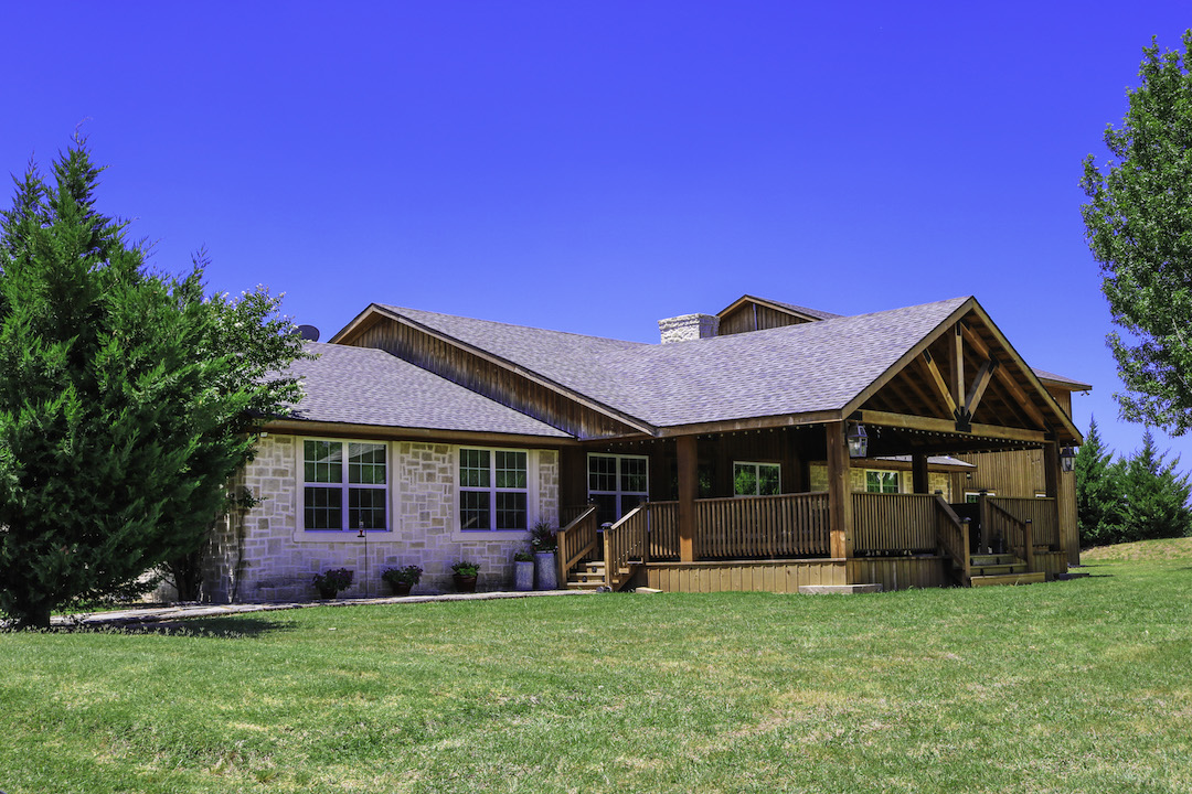 A single-story farmhouse with a prominent wooden gable, front deck and green lawn under a clear blue sky.