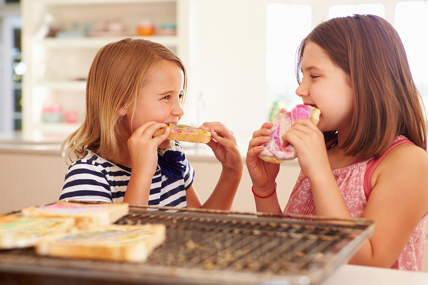 Two girls smiling, enjoying colorful frosted toasts in a bright kitchen, with a wire rack and unfocused background.