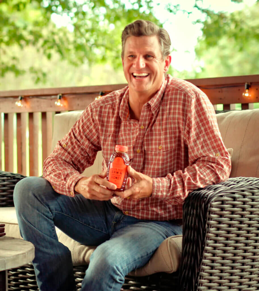 The man Nature Nate smiling, holding a bottle of Nate’s honey on a porch.