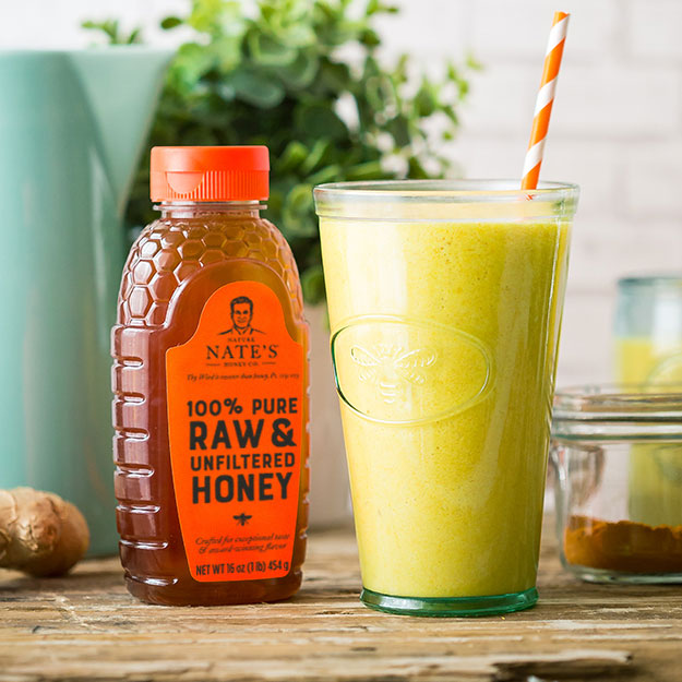 Green turmeric and ginger smoothie with a straw next to a bottle of Nate's raw honey on a wooden table.