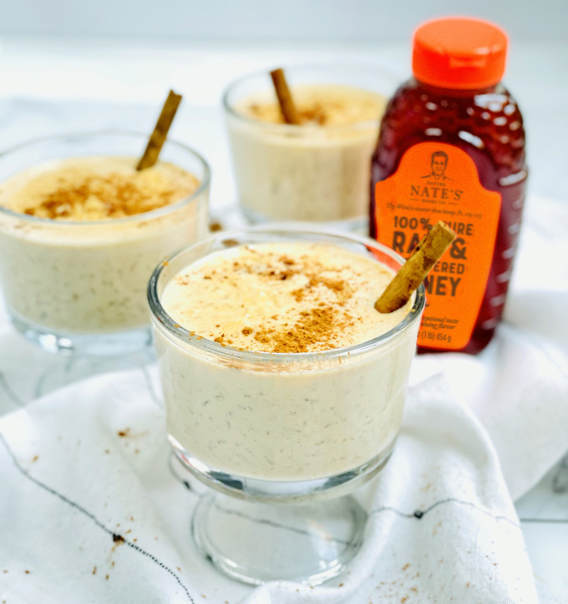 Creamy rice pudding in glasses with cinnamon, a bottle of Nate’s honey with an orange cap in the backdrop.
