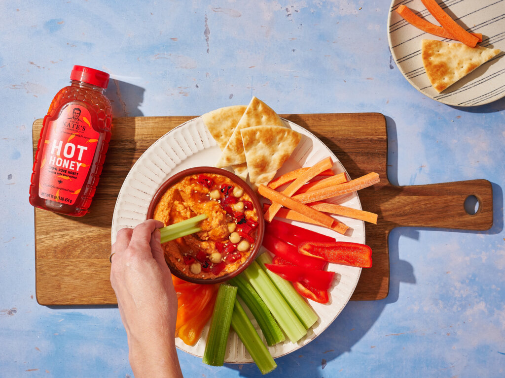 Hand dipping celery in hummus with veggies, pita, 
Nate’s hot honey bottle, all on a wooden board.