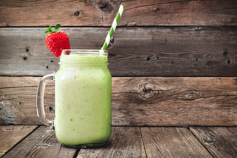 A fresh green veggie smoothie in a mason jar with a straw, topped with a strawberry, on a rustic wooden background.