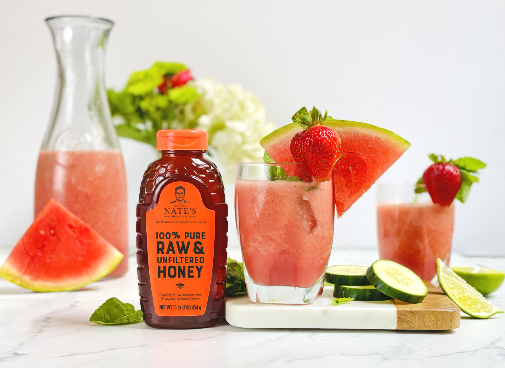 Watermelon slushies with lime, a bottle of Nate’s honey, and watermelon and cucumber slices on a marble counter.