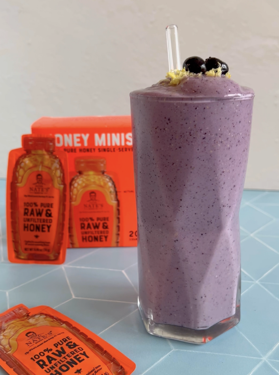 A purple blueberry smoothie garnished with blueberries, next to Nature Nate's honey Minis.