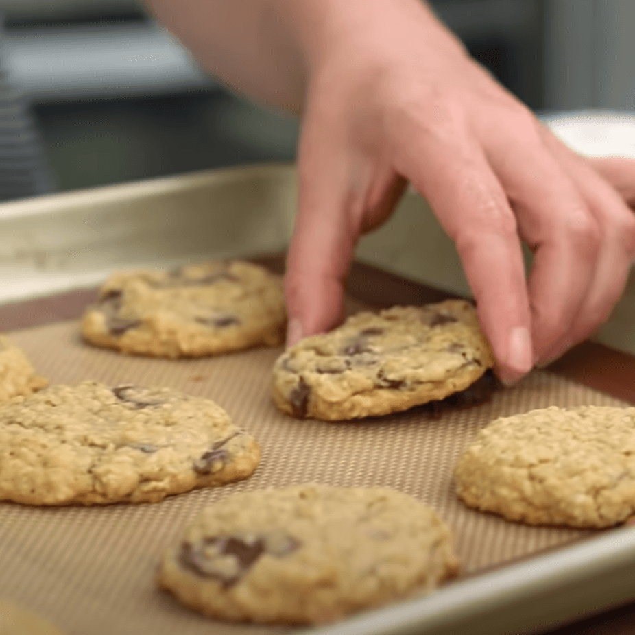 A hand reaching for chocolate chip oatmeal cookies on a baking sheet.