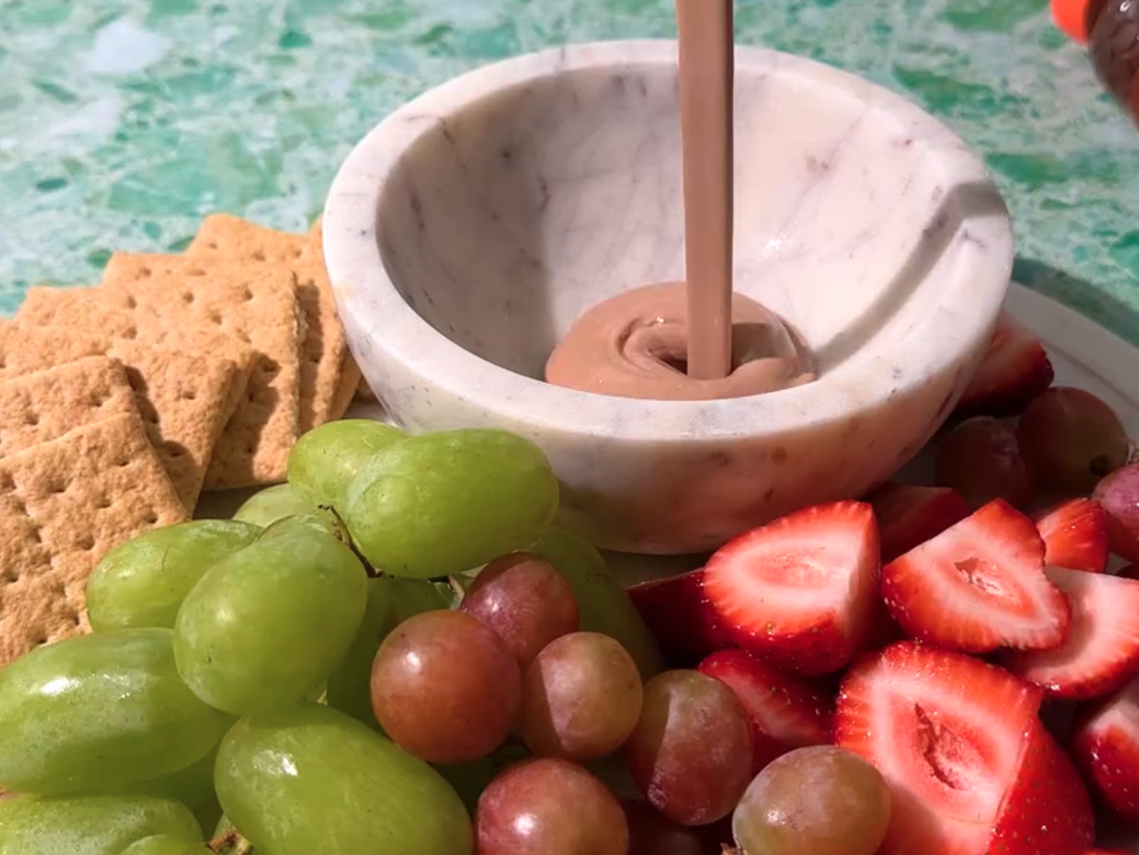 Chocolate dip made with Nate’s honey in a marble bowl surrounded by fruits and graham crackers on a marbled surface.