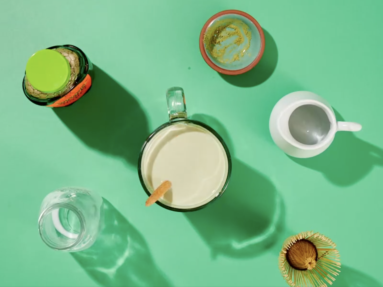 Matcha latte in the center with match powder, hot water, hot milk and Nate's organic honey surrounding it on a green background.