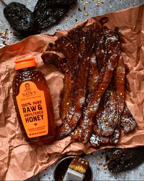 Honey-glazed bacon and a bottle of Nate's raw & unfiltered honey with an orange cap on a speckled counter.