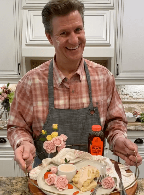 The man Nature Nate in a plaid shirt and apron, holding a tray, stands in a kitchen with a bottle of Nate's honey.