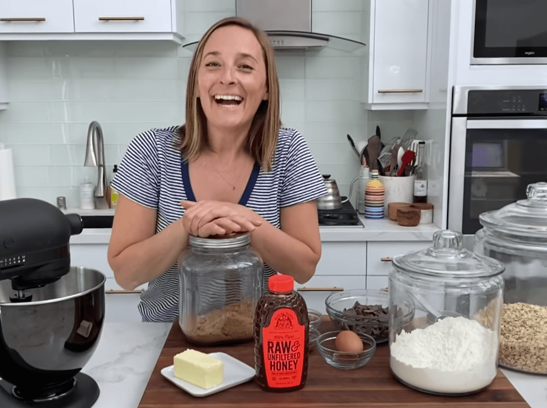 A woman smiling in a kitchen with baking  ingredients and a bottle of Nate's honey.