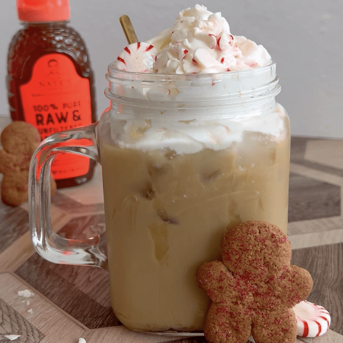 Peppermint honey latte in a mug topped with whipped cream. Displayed with peppermint candies, gingerbread cookies and Nate’s honey bottle.