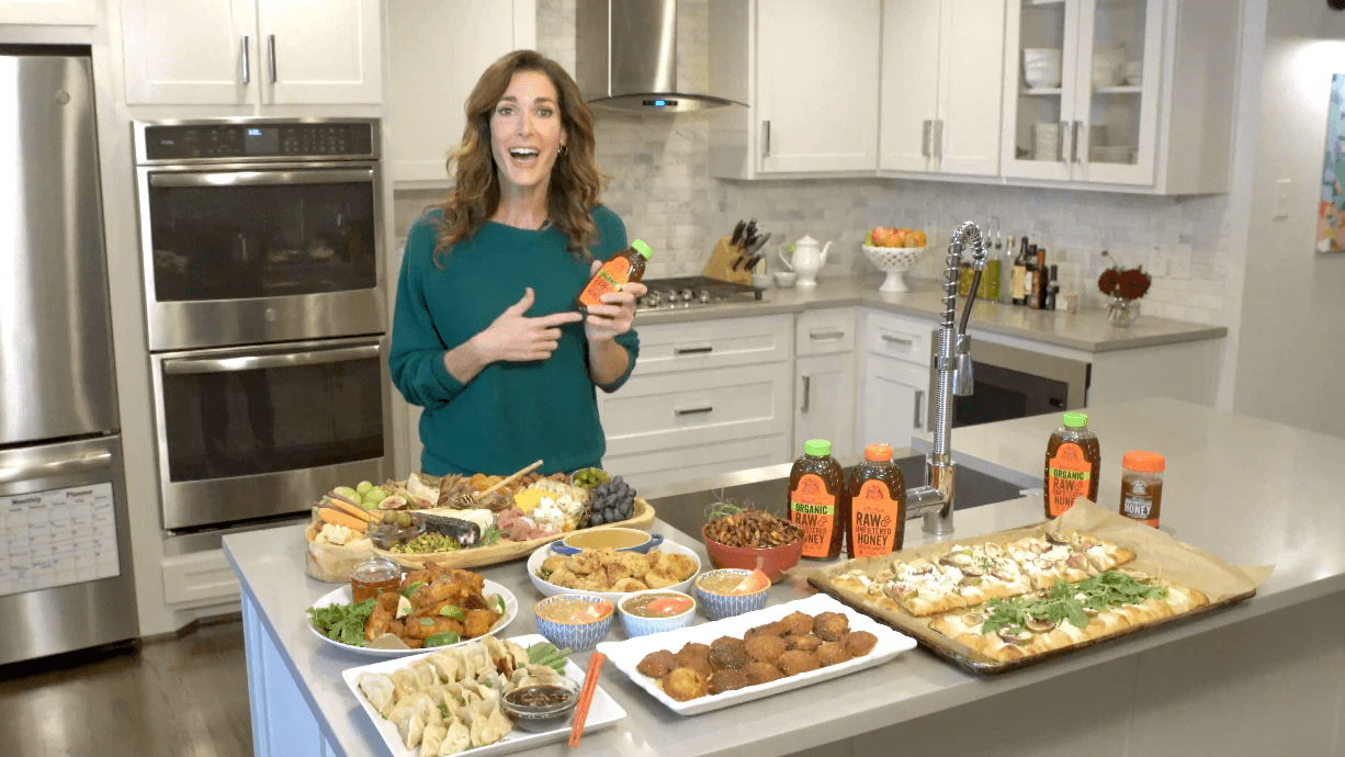 A smiling woman presenting various dishes and Nate’s honey bottles on a kitchen island.