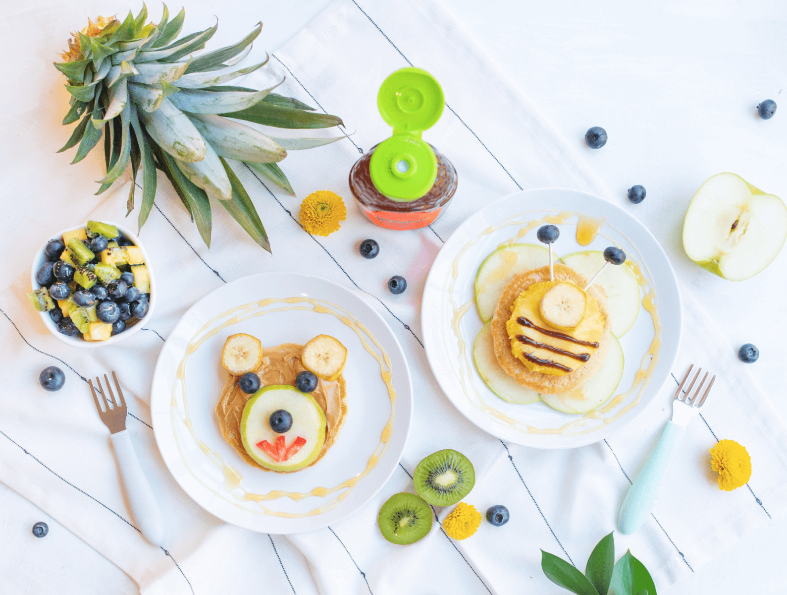 Pancake breakfast with pancakes shaped like animals and fruit, and Nate's organic honey bottle on a white table.