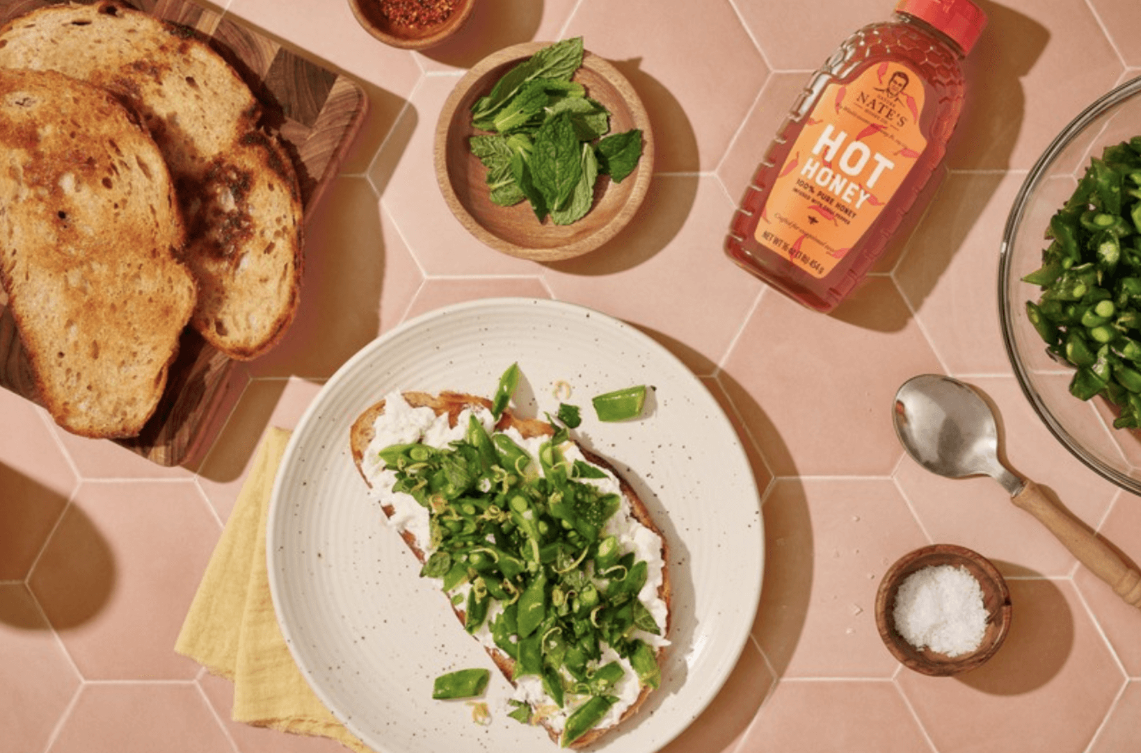 Burrata and snap pea toast on a plate, a bowl of snap peas, mint leaves and a bottle of Nate's hot honey on a pink table.