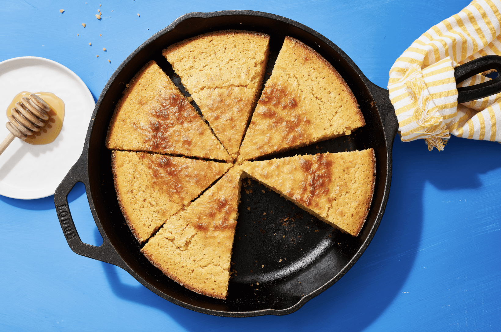 A skillet of golden-brown cornbread cut into wedges on a blue surface with honey and a cloth napkin alongside.