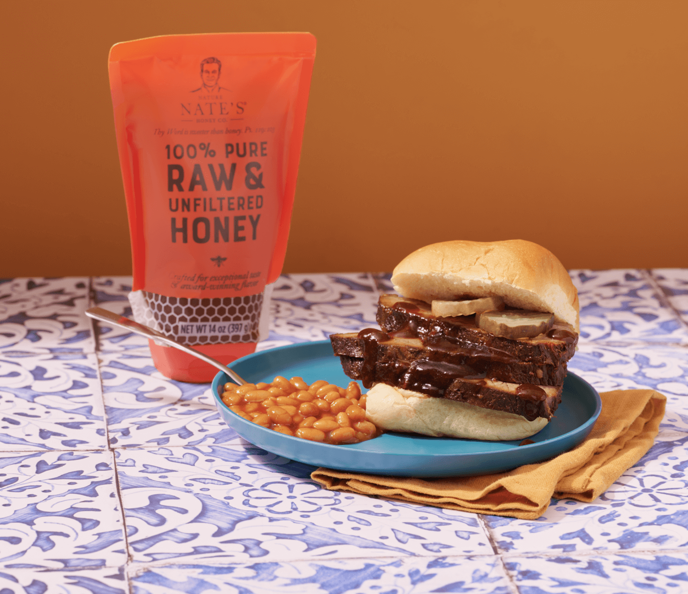 A barbecue brisket sandwich on a blue plate with baked beans, a napkin and a pouch of Nate's raw & unfiltered honey.