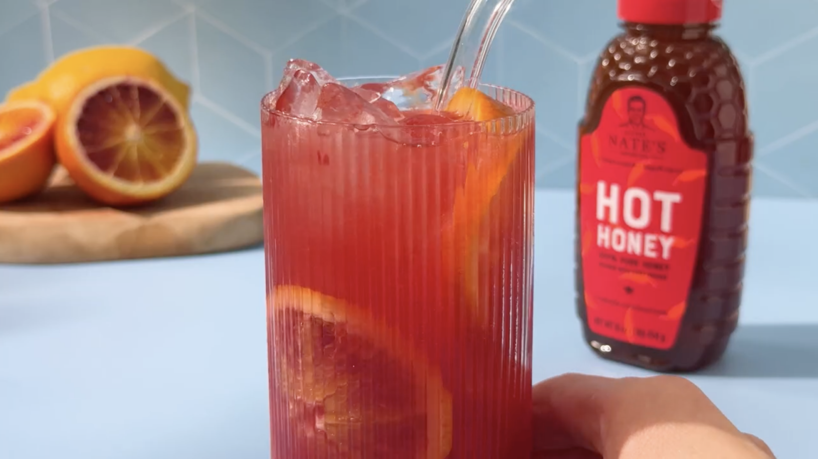 Glass of blood orange cocktail with ice and orange slices and Nate's hot honey bottle in the background.