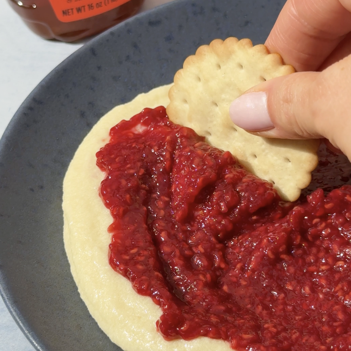 Cracker being dipped in Whipped Brie Dip with Honey Raspberries, with a bottle of Nate's organic honey in the background.