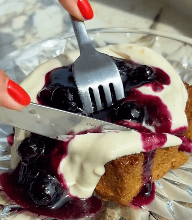 Honey butter toast topped with cream and blueberry compote being cut with a fork and knife.