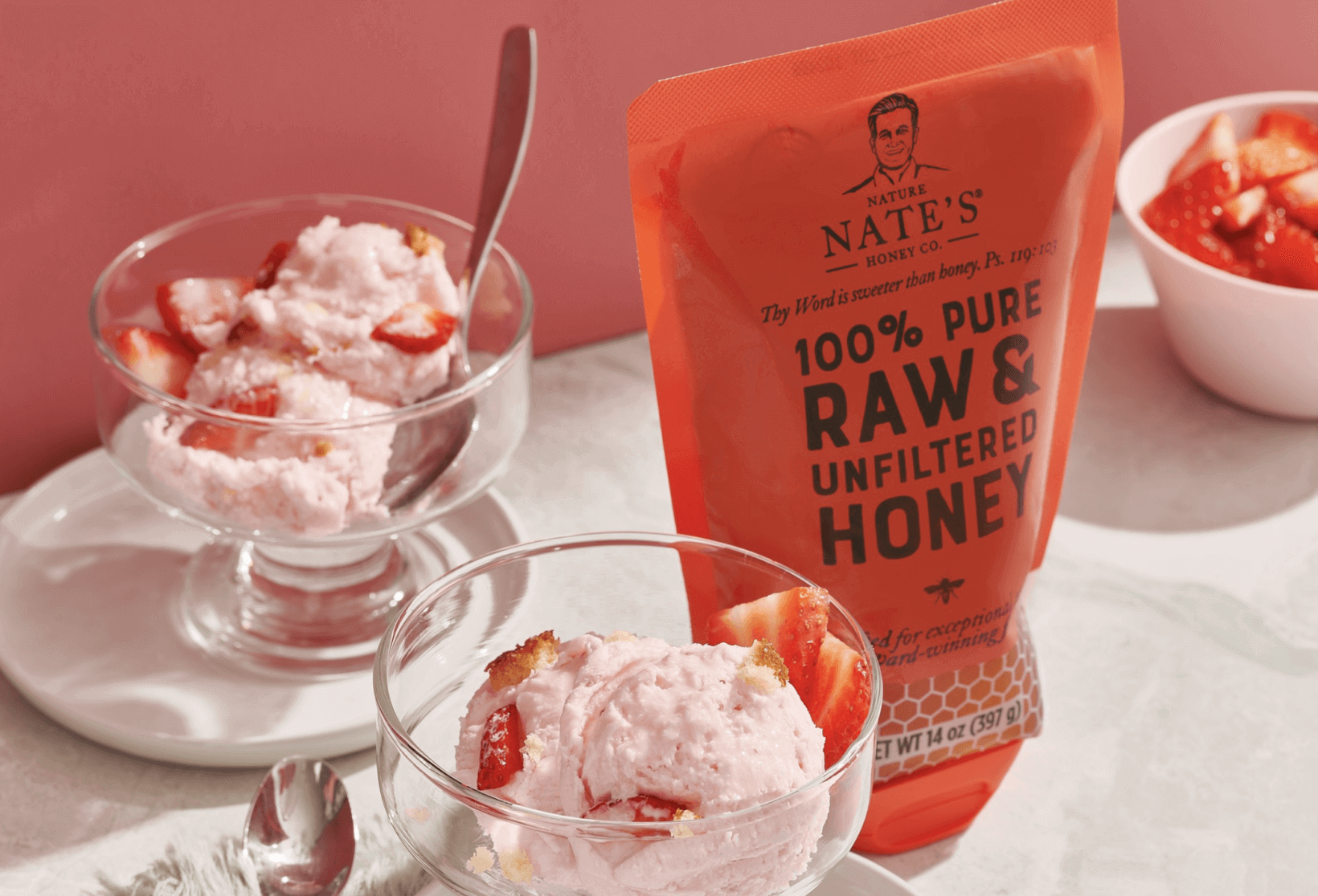 A pouch of Nates 100% pure raw & unfiltered honey next to bowls of strawberry ice cream on a table with a pink hue.