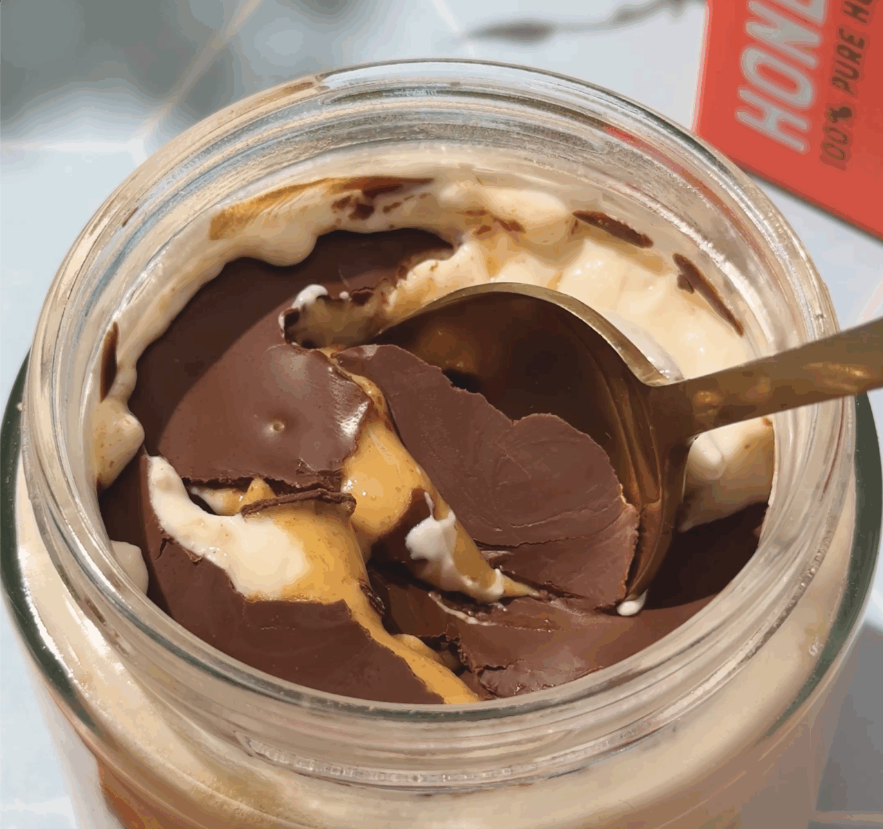Spoon cracking into a chocolate shell topping over creamy yogurt, peanut butter, and honey in a glass jar.